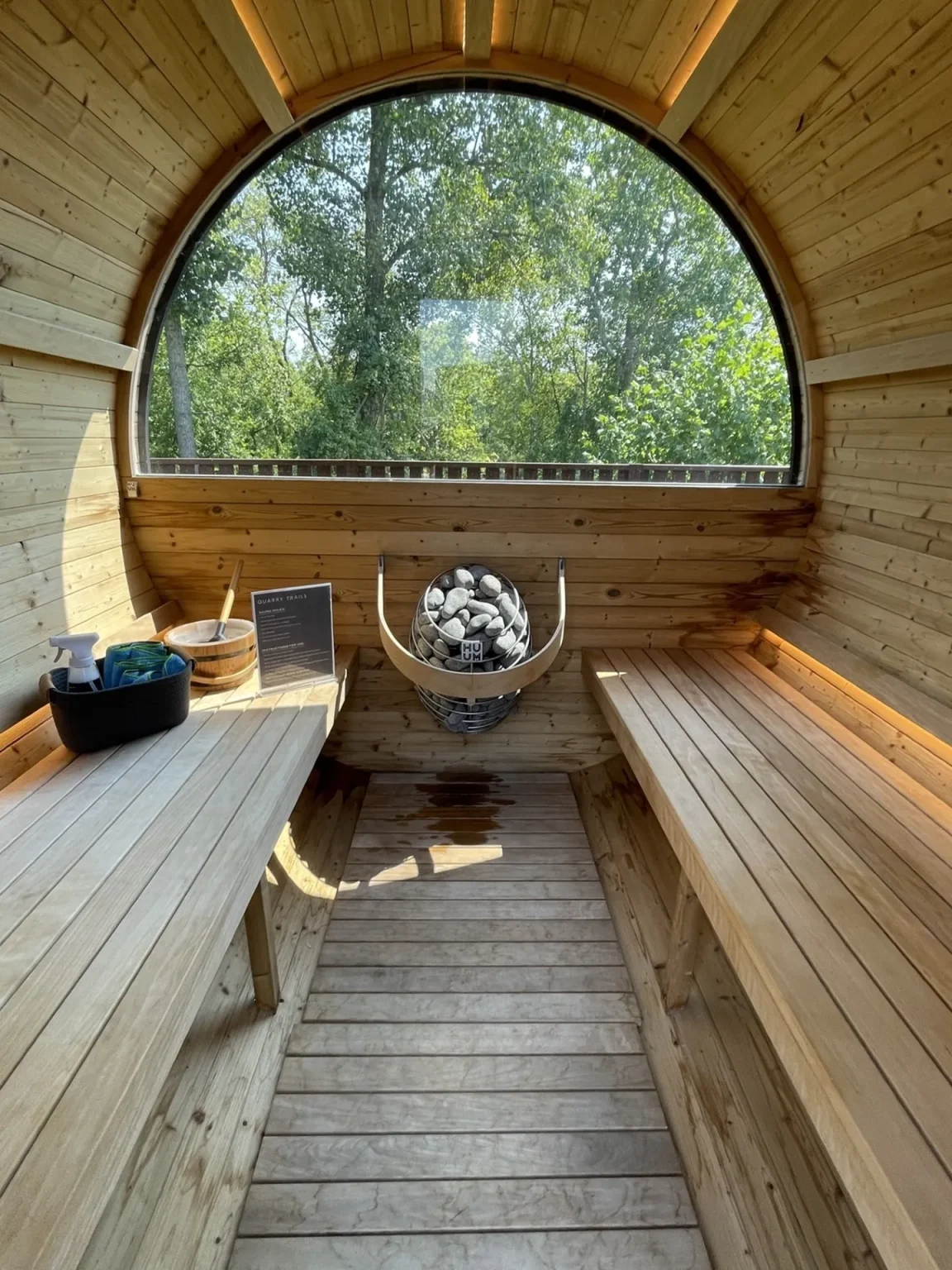 Interior of a wooden sauna with benches on both sides, a central heater with stones, and a large arched window showing trees outside.