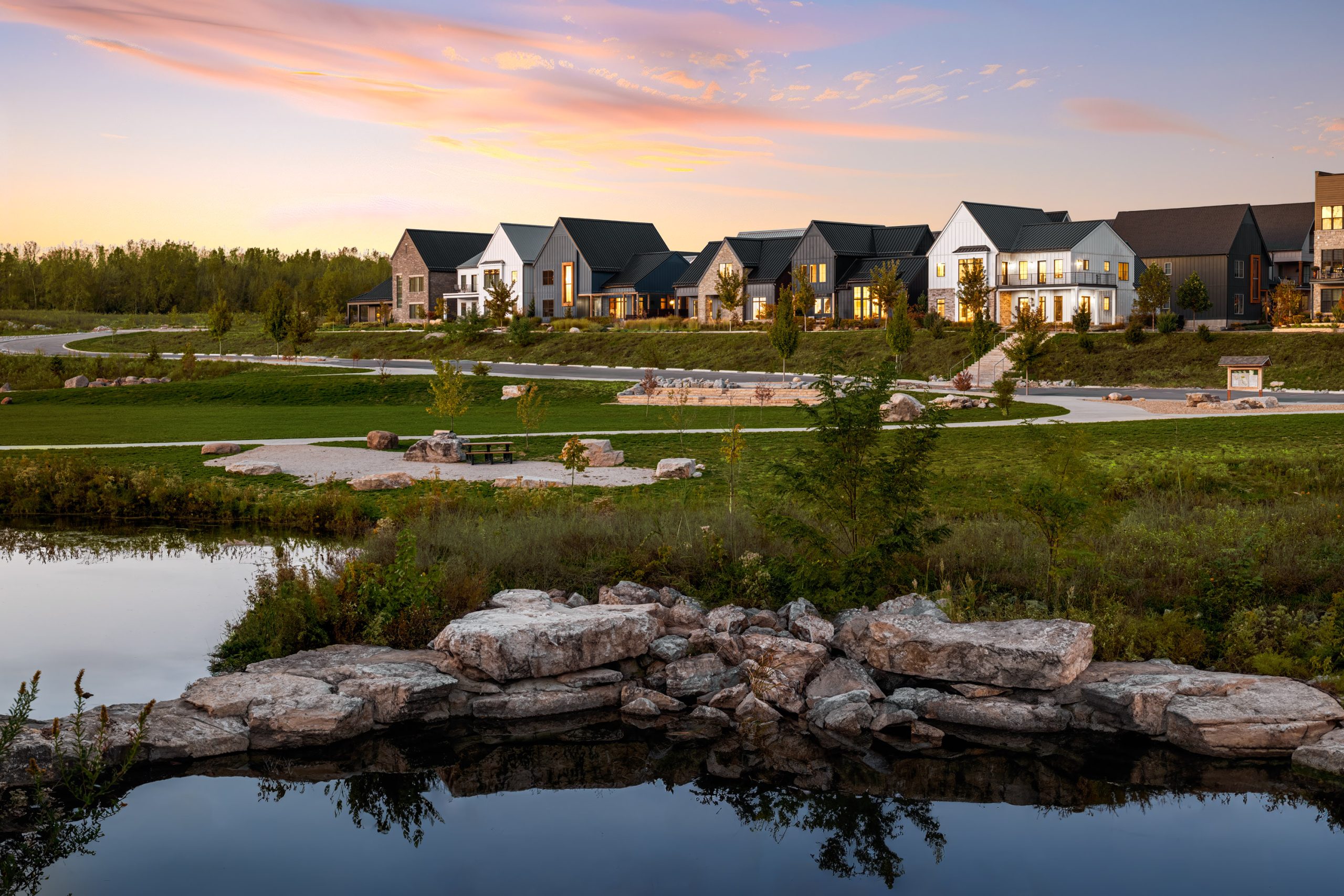A row of modern houses with large windows, developed by Thrive Companies, is set against a sunset sky. In the foreground, theres a pond surrounded by rocks and greenery, reflecting the elegance of this exceptional real estate.