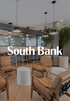 Modern office lounge area with brown leather chairs, small white tables, hanging pendant lights, and potted plants. The words South Bank at Quarry Trails are centered over the image.