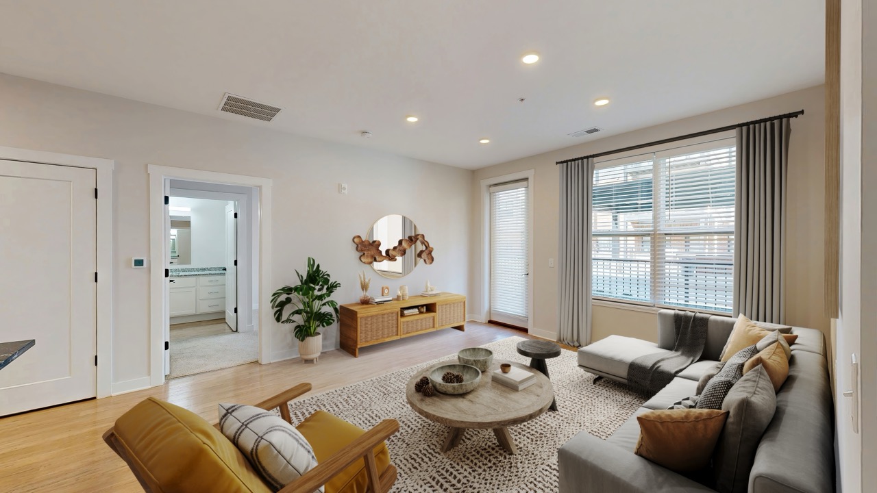 A modern living room at Trace Quarter features a gray sectional sofa, yellow armchairs, a round coffee table, a wooden TV console, and large windows with curtains letting in abundant natural light.