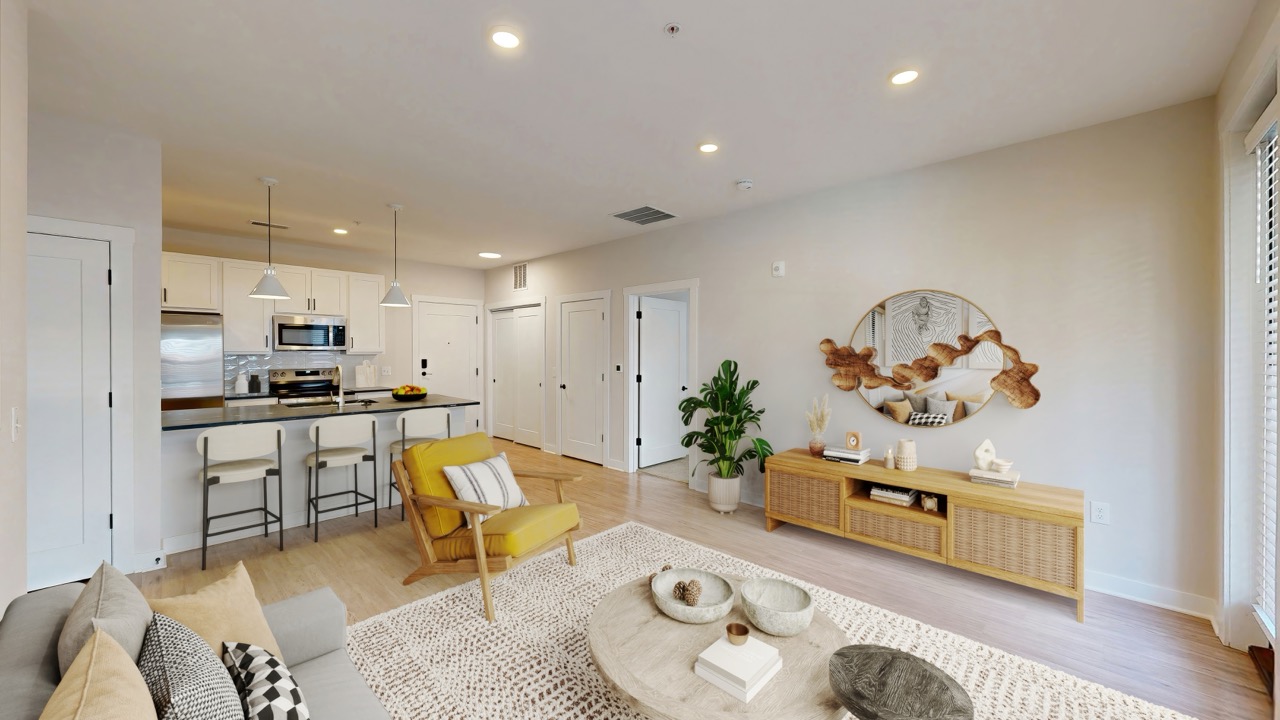 Modern living room at Trace Quarter with a neutral color palette, featuring a yellow chair, sofa, round coffee table, TV console, wall art, and an open kitchen with barstools in the background.