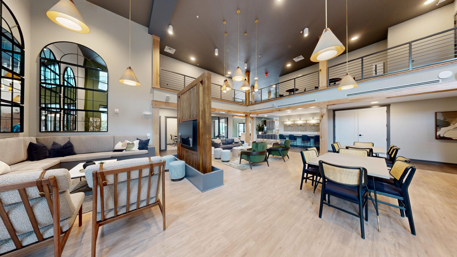 Modern, spacious Trace Quarter lounge area with wooden flooring, mixed seating, a central divider featuring plants and a TV, high ceilings, pendant lighting, and a mezzanine level with railing.