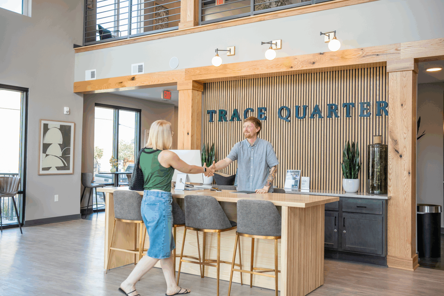 A woman and a man shake hands at a reception desk in a modern, well-lit Trace Quarter office, with the distinctive TRACE QUARTER signage on the wall behind them.