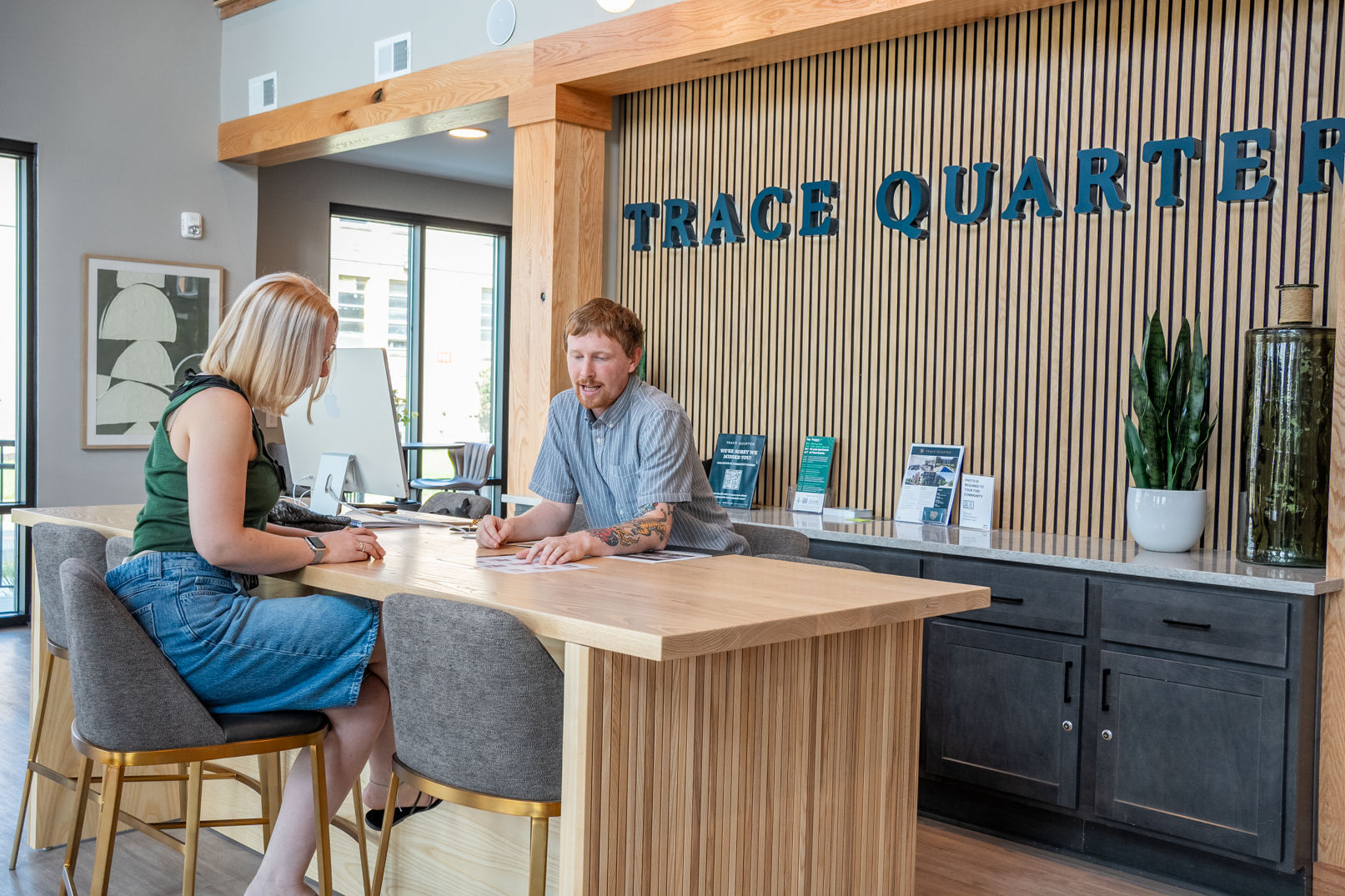A man and a woman sit at a wooden desk in a modern office, discussing documents beneath the TRACE QUARTER sign. The professional setting highlights their focus on Trace Quarters business strategies.
