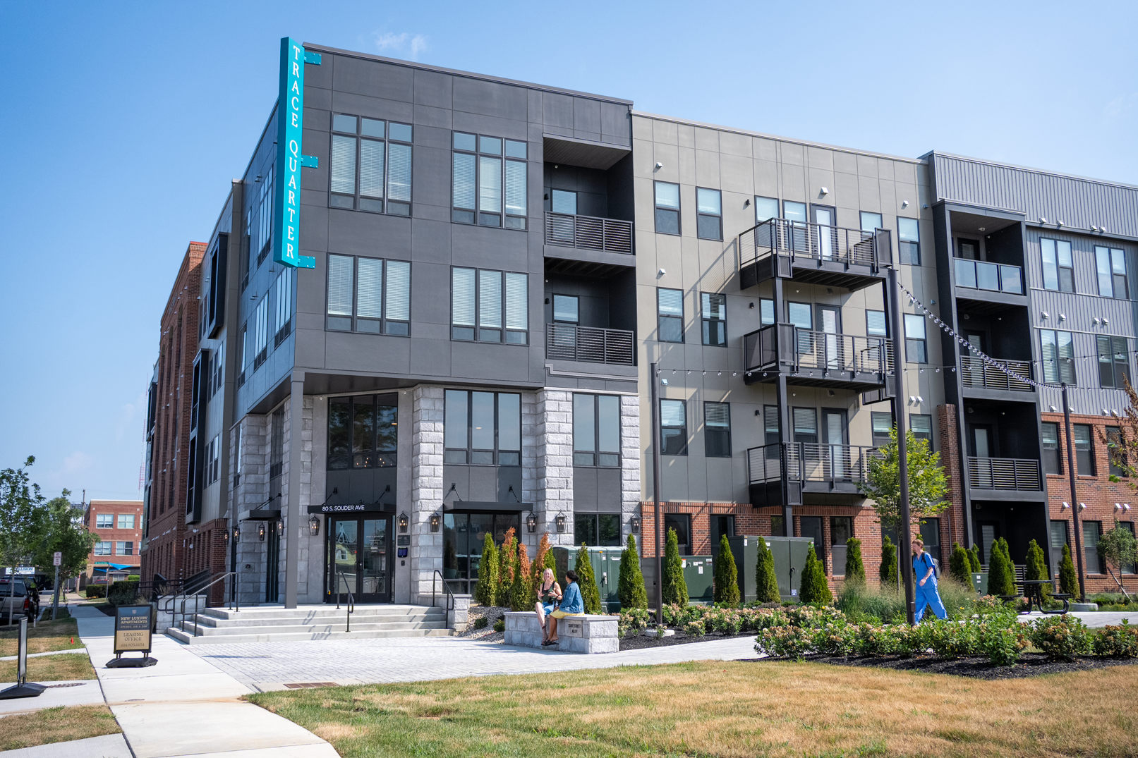 Modern apartment building with gray and brick exterior, balconies, and a sign reading PLACE ON 4TH, located near Trace Quarter. Landscaped entrance and people walking nearby on a sunny day.
