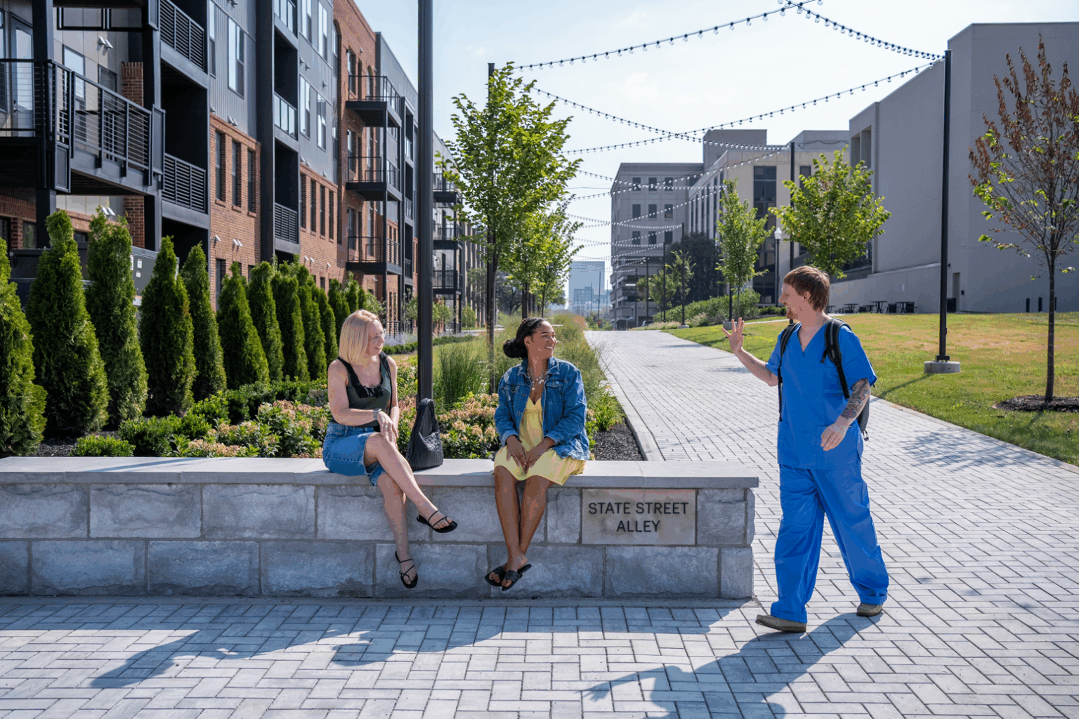 Two women sit and talk on a low stone wall labeled “State Street Alley” in the Trace Quarter, while a person in blue scrubs waves as they walk by on a paved path in this urban area.