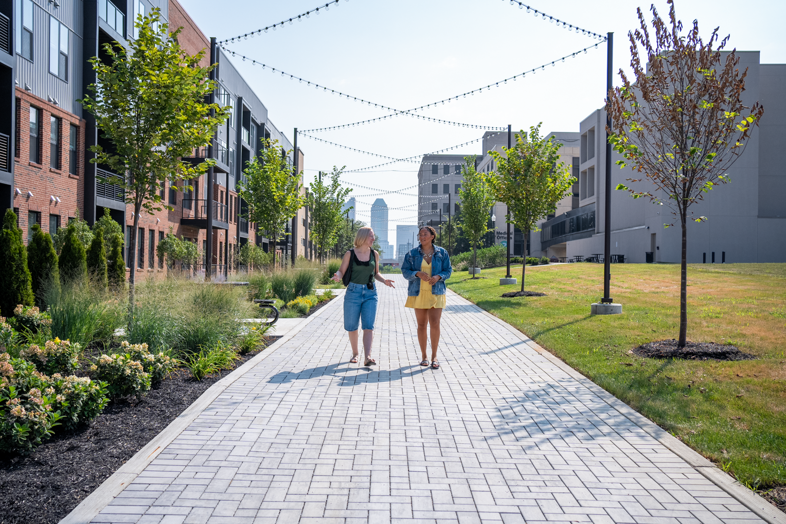 Two people stroll along a paved path lined with trees and modern buildings in Trace Quarter on a sunny day. String lights are hung overhead along the walkway.