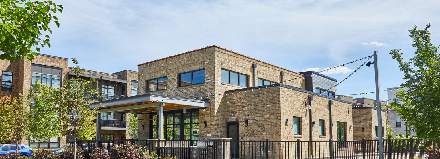 Modern brick building with large windows, a metal canopy, and a fenced yard, surrounded by trees on a clear day.