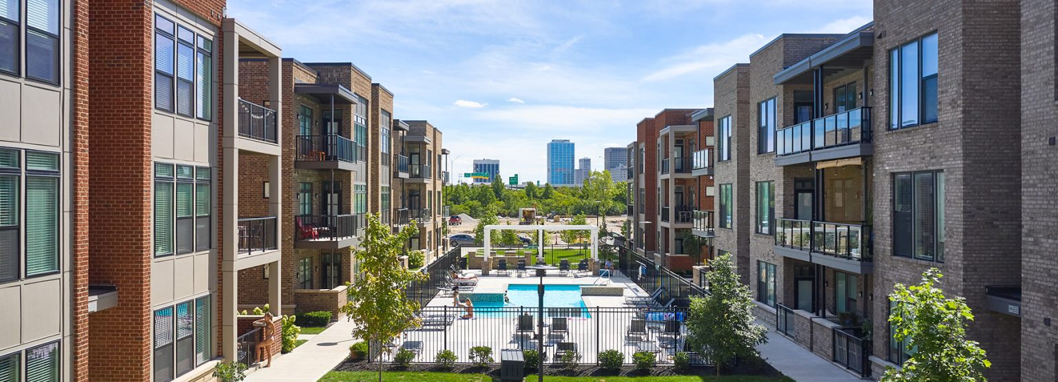Modern apartment complex with balconies, centered courtyard, and a pool. City skyline visible in the background under a clear blue sky.