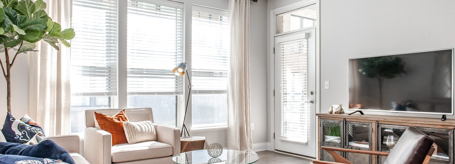 Bright living room with large windows, beige armchair, TV, wooden console, and glass coffee table. A floor lamp and indoor plant add decor. Light curtains cover the windows.