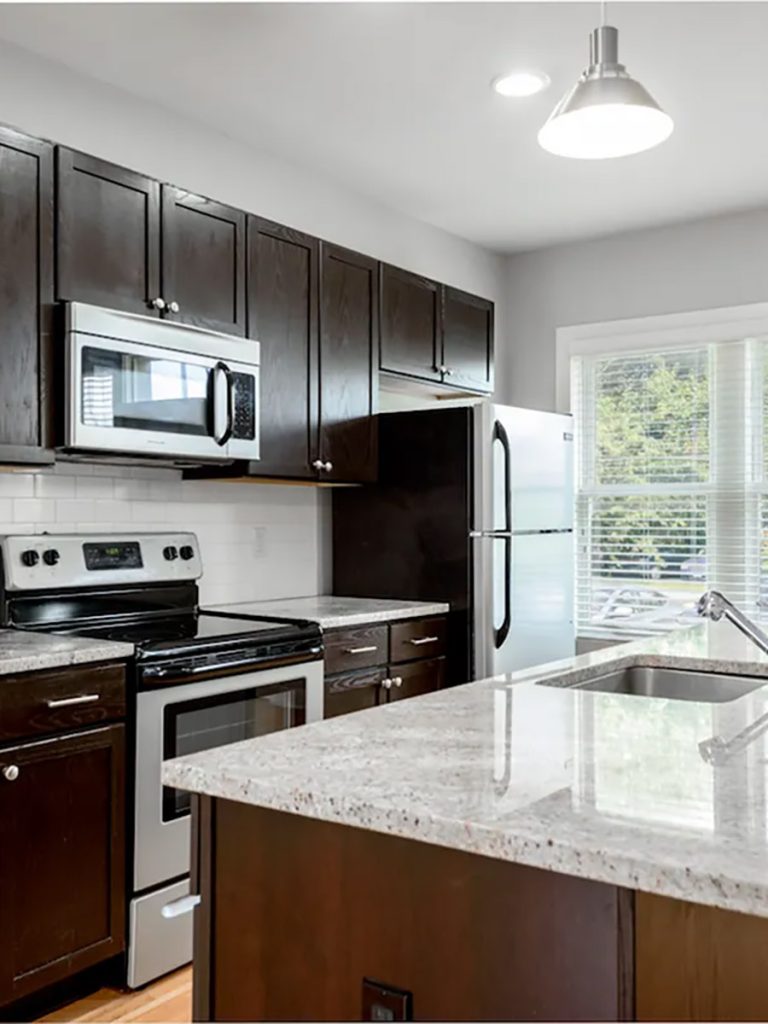 Modern kitchen with dark wood cabinets, stainless steel appliances, and a granite countertop island. Large window to the right providing natural light.