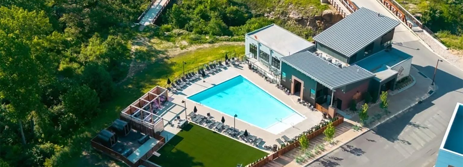 Aerial view of a recreational area with a swimming pool, lounge chairs, buildings, an outdoor seating area, and surrounding greenery.