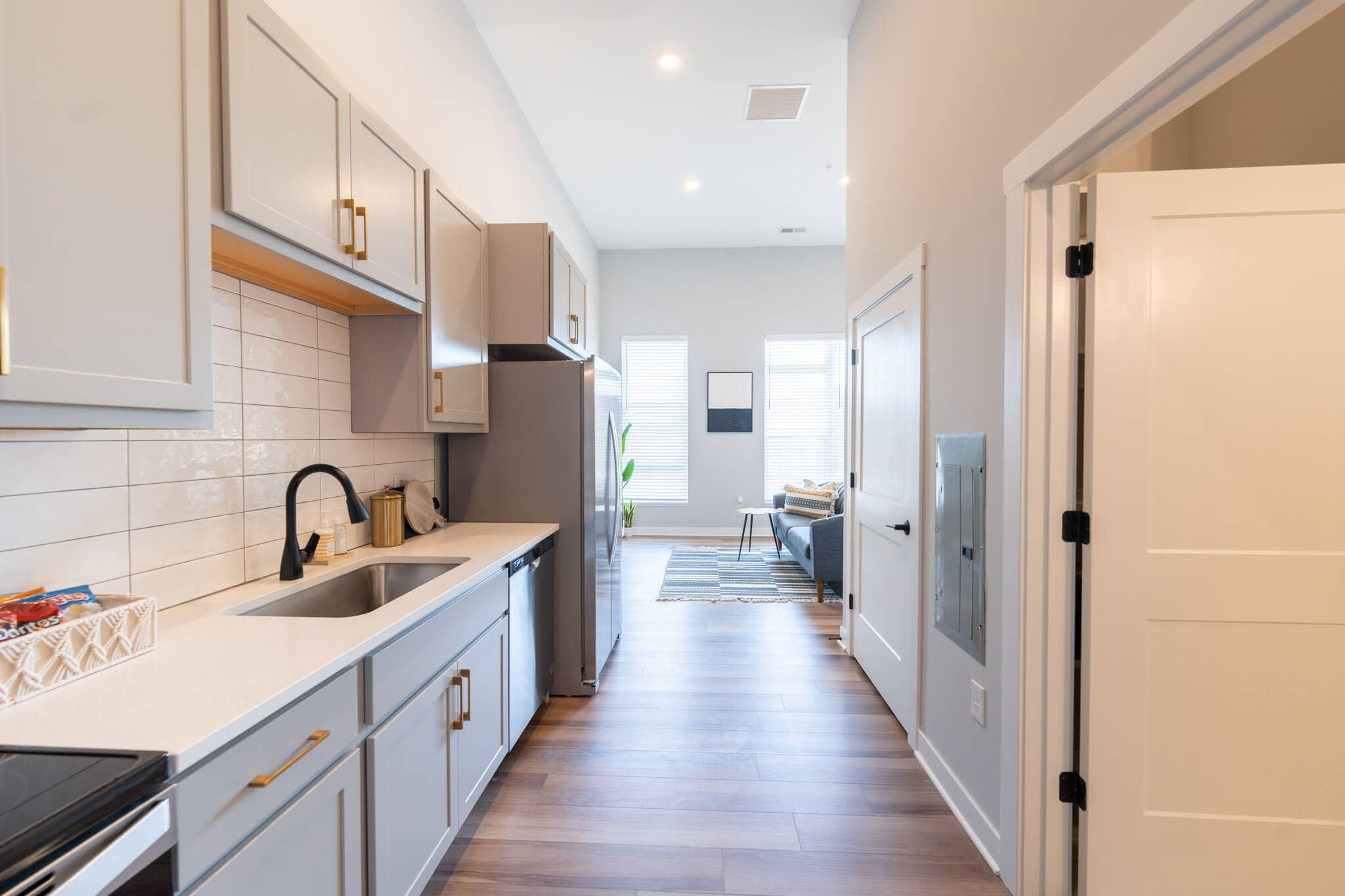 Modern galley kitchen with light gray cabinets, white countertops, stainless steel appliances, and a view into a bright living area with large windows.