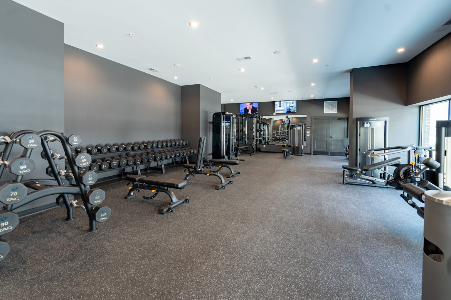 Modern gym with free weights, benches, weight machines, and cardio equipment in a spacious room with gray walls and bright lighting.