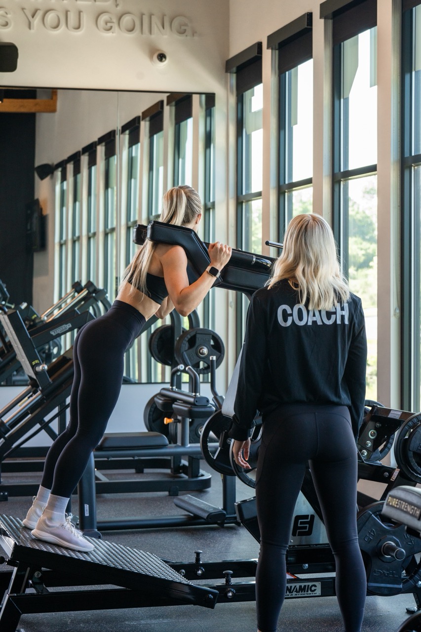 A woman performs a squat exercise on gym equipment while another woman, wearing a COACH jacket, observes and provides guidance.