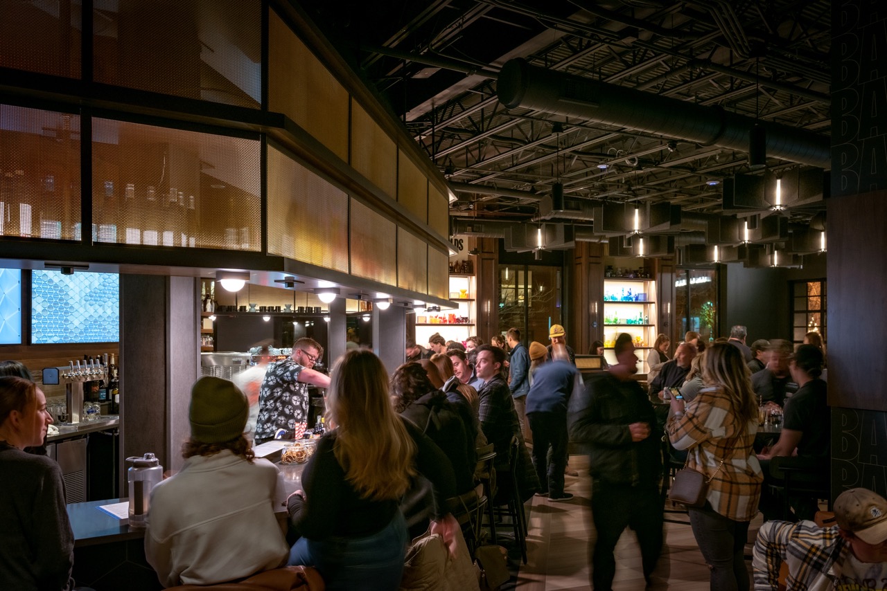 Busy bar scene with people seated and standing, dim lighting, and shelves of bottles. A bartender serves drinks while patrons socialize.