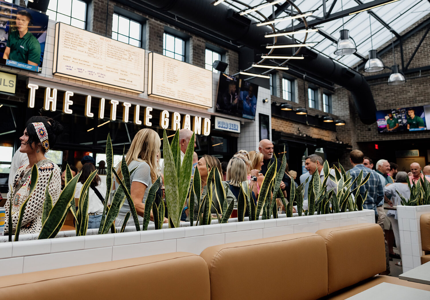 A group of people socializing inside The Little Grand, a Thrive Companies real estate gem, adorned with plants, cushioned seating, and bright signage. Menu boards and TVs are visible in the background.