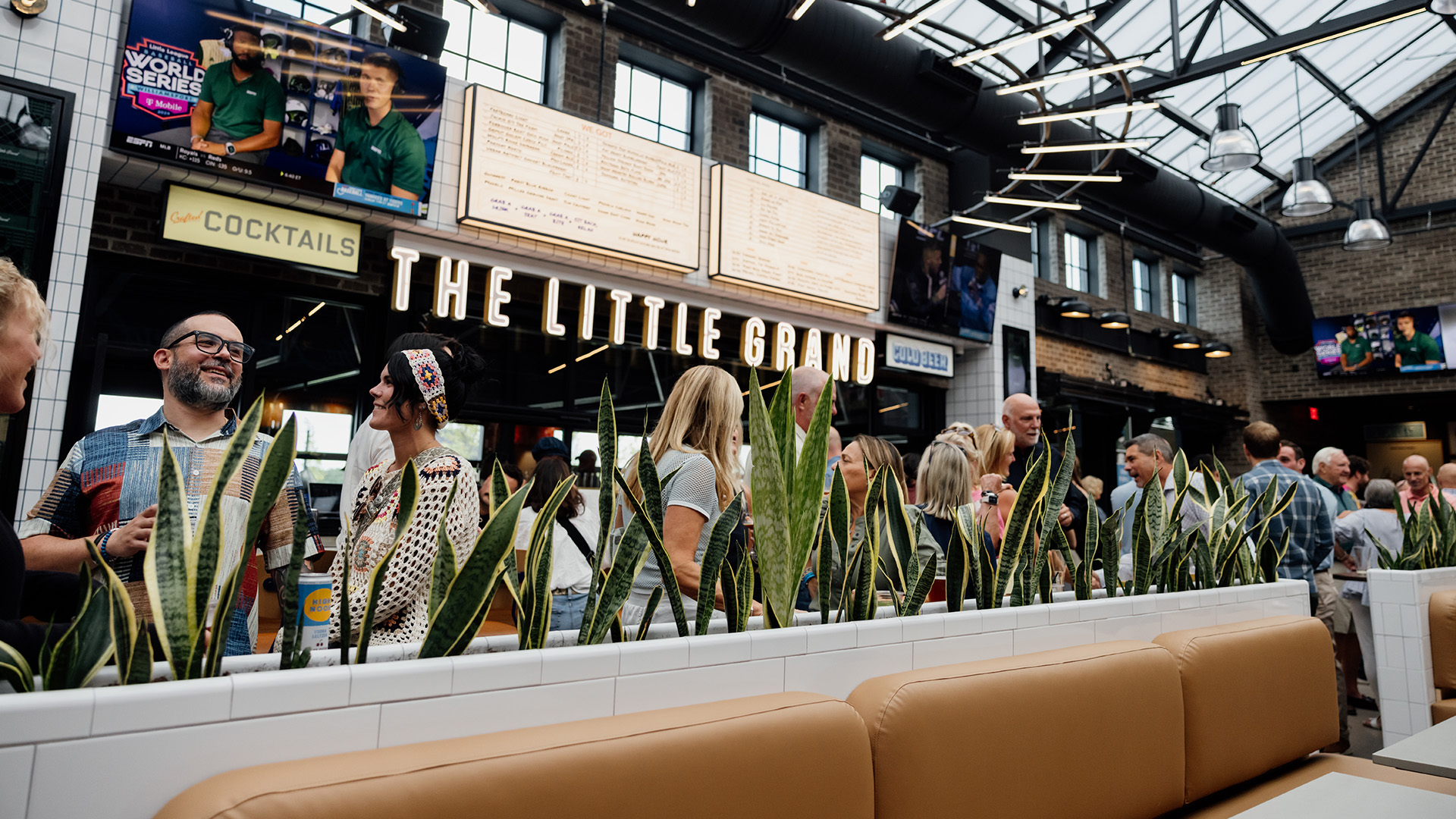 A group of people socializing inside The Little Grand, a Thrive Companies real estate gem, adorned with plants, cushioned seating, and bright signage. Menu boards and TVs are visible in the background.