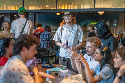 A group of people socializing at a bar, with one person standing and smiling while holding a drink. Various drinks and personal items are on the table.