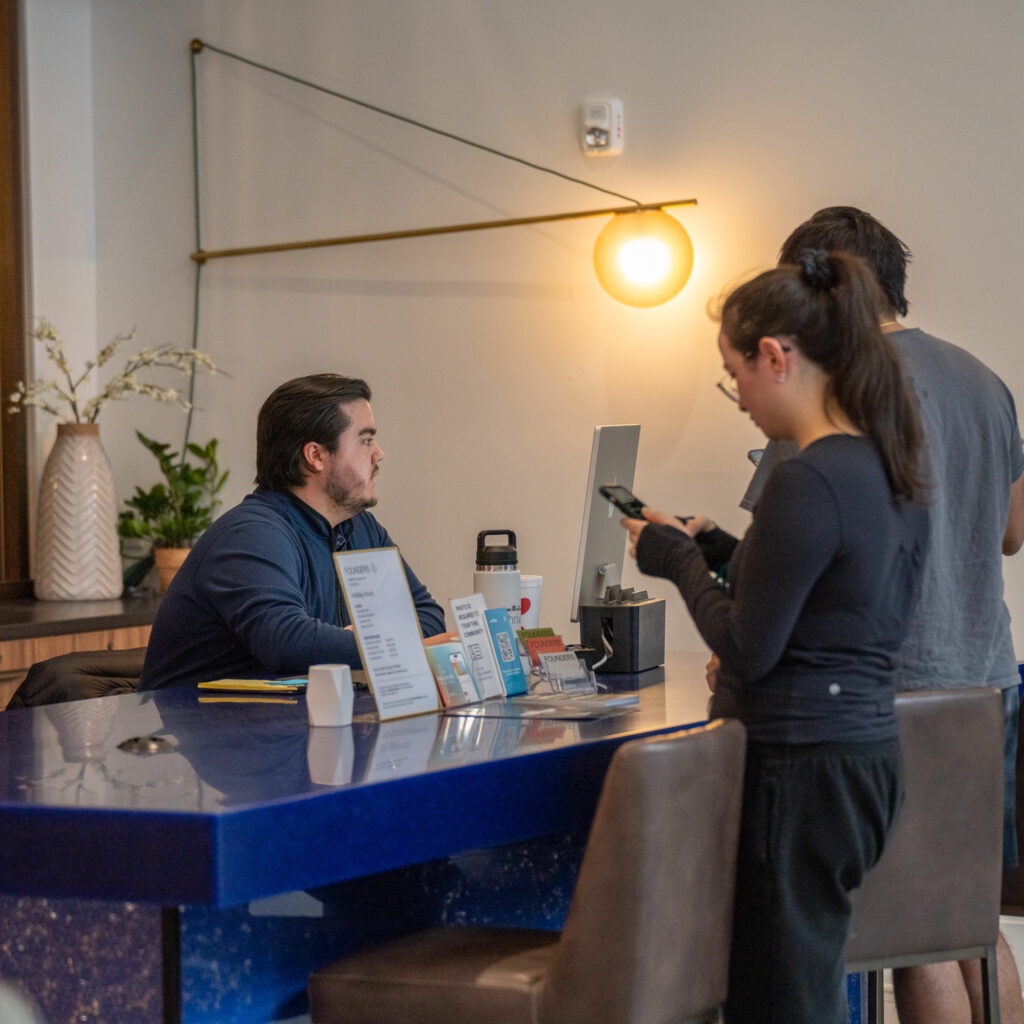 A man sits at a blue reception desk using a computer while two people stand on the other side, one looking at a phone. A modern light fixture and vase are in the background.