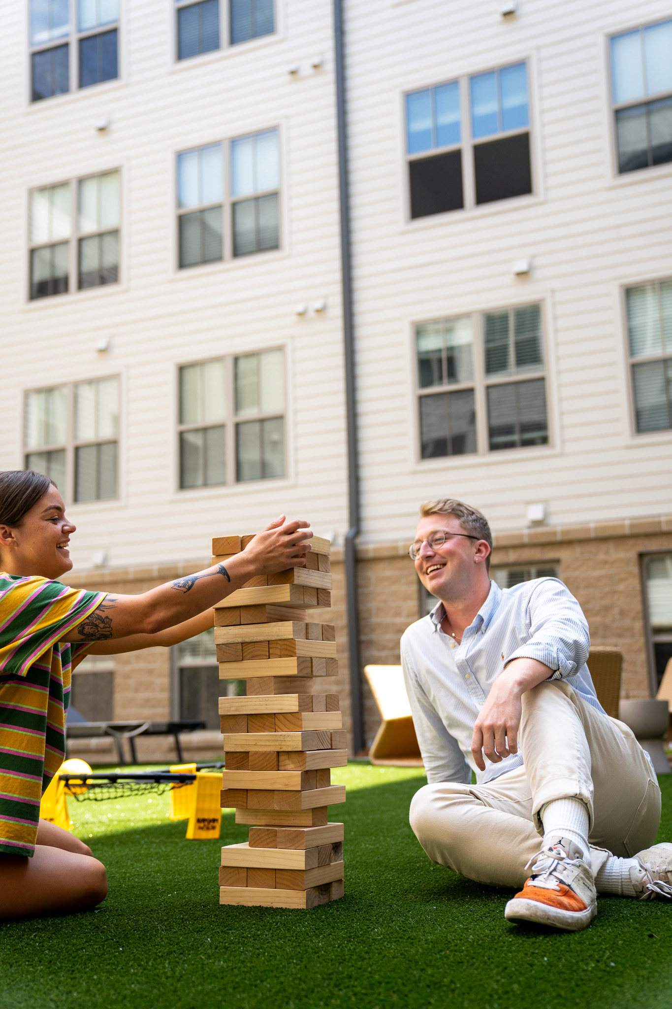 Two people sit on artificial grass in a courtyard playing giant Jenga, with apartment building windows in the background.