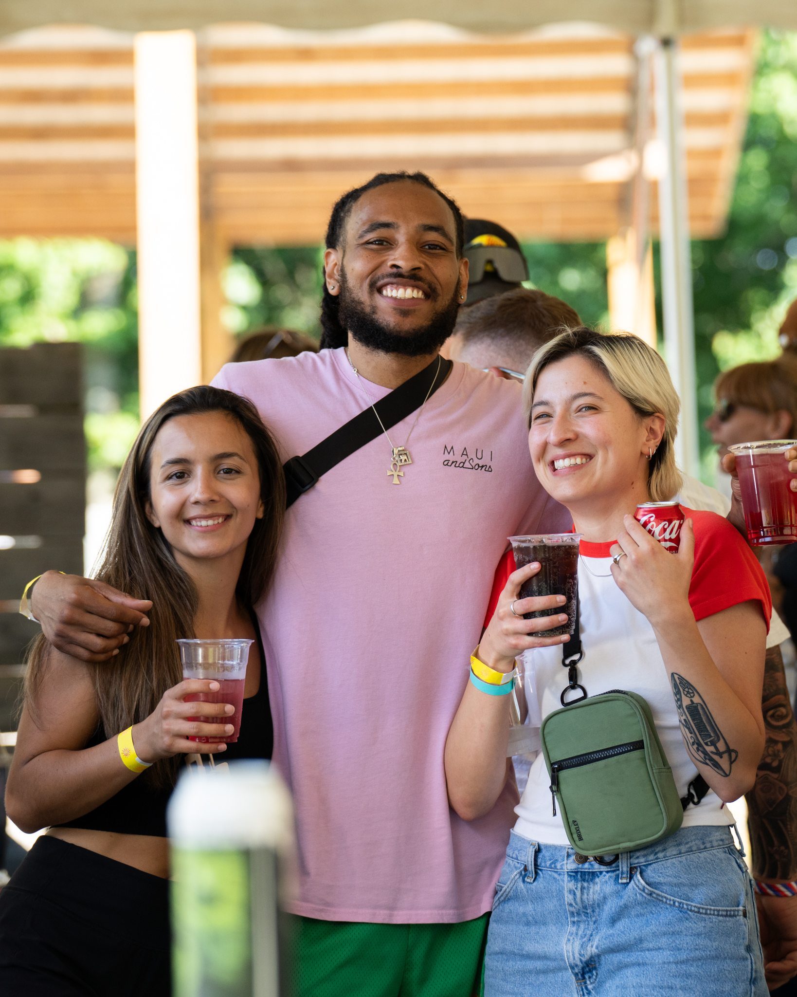 Three people stand close together outdoors, smiling and holding drinks, with wristbands visible, under a tent with sunlight in the background.