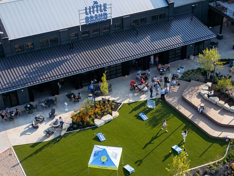 Aerial view of a grassy area with people playing cornhole outside a building named The Little Grand. There are tables, chairs, and landscaping surrounding the playing area.