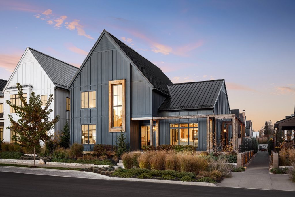 Modern farmhouse with steel siding, large windows, and a pitched roof. Front yard has landscaped plants and a sidewalk. Sky is clear at dusk.