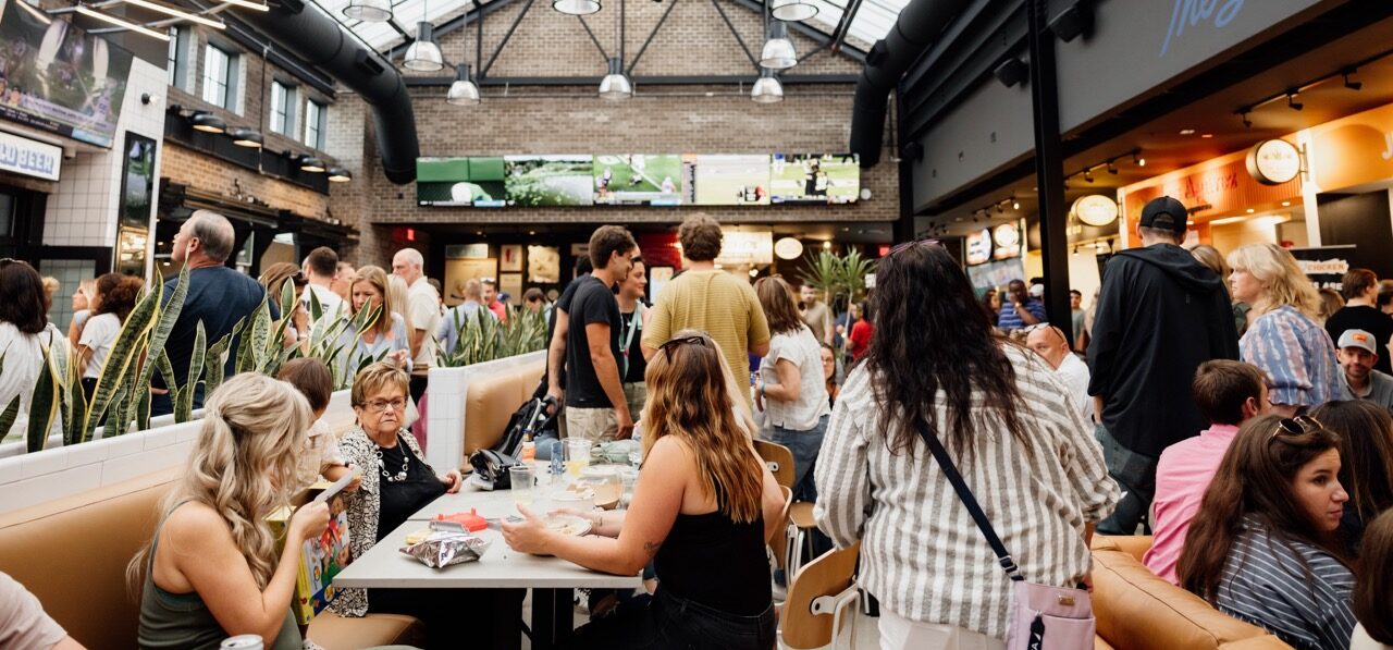 People sitting and standing inside a busy food court with a glass ceiling, restaurant signage, and multiple television screens displaying sports.