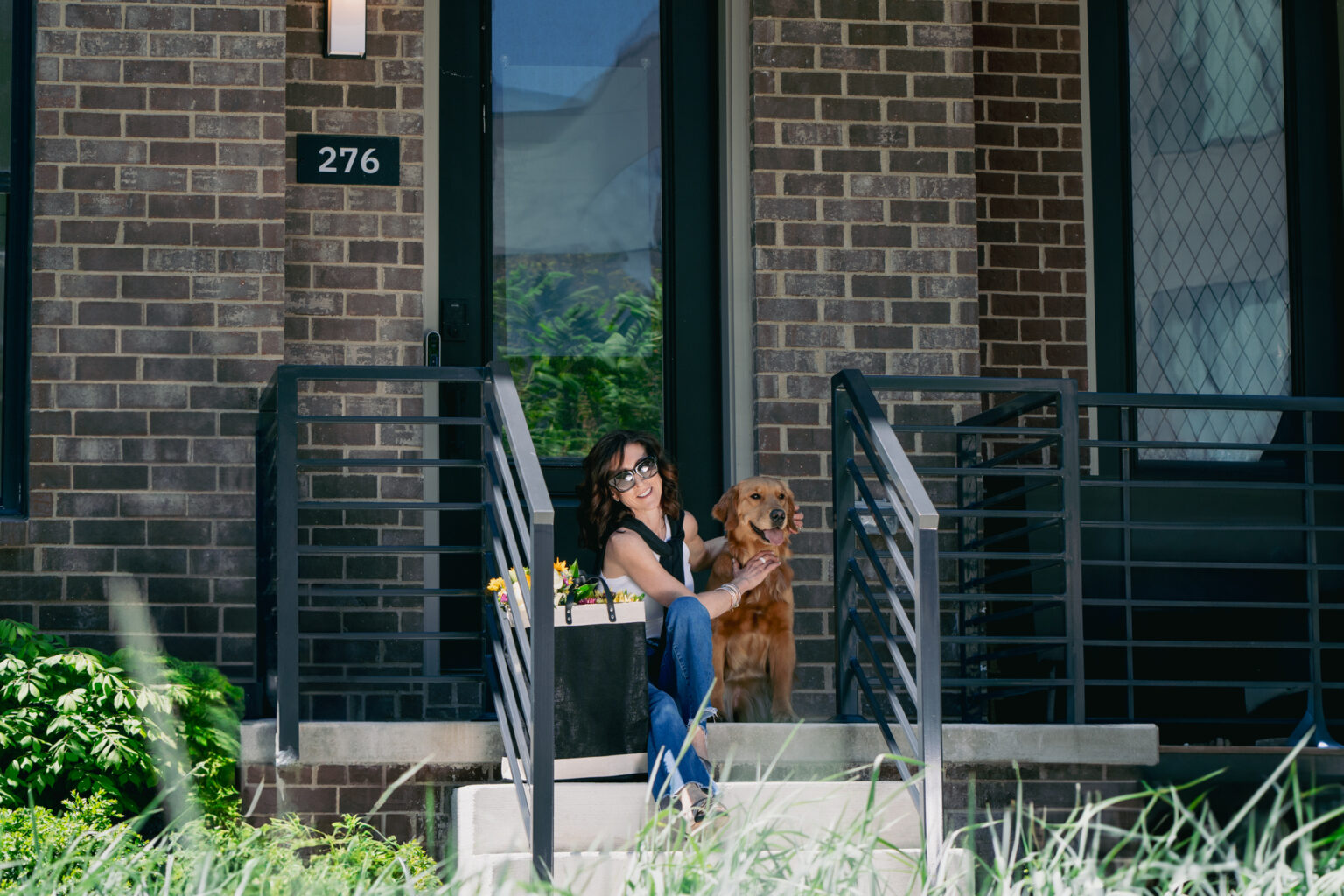 A woman sits on the front steps of a brick building at address 276, smiling and holding a golden retriever dog.