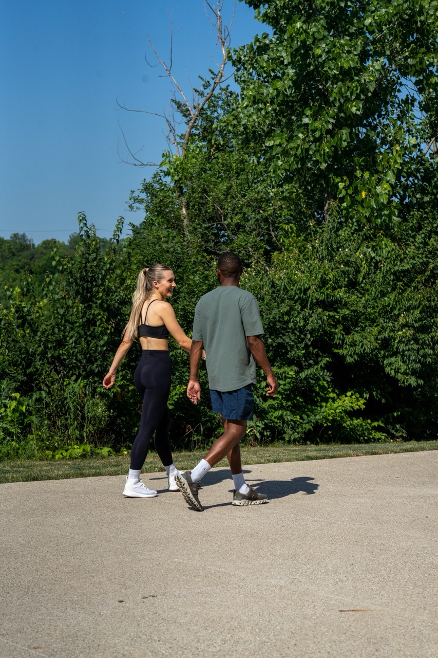 A woman and a man walk together on a paved path outside, surrounded by green trees and bushes on a sunny day.