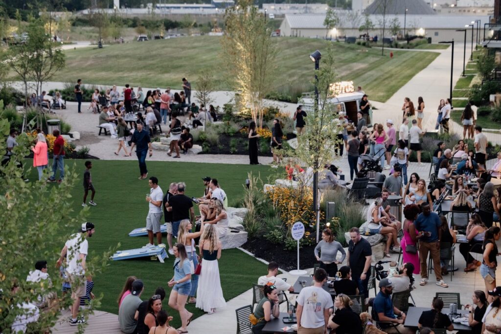 Outdoor gathering in a park with people socializing, sitting on grass and benches, and playing games. Trees and pathways are visible in the background.