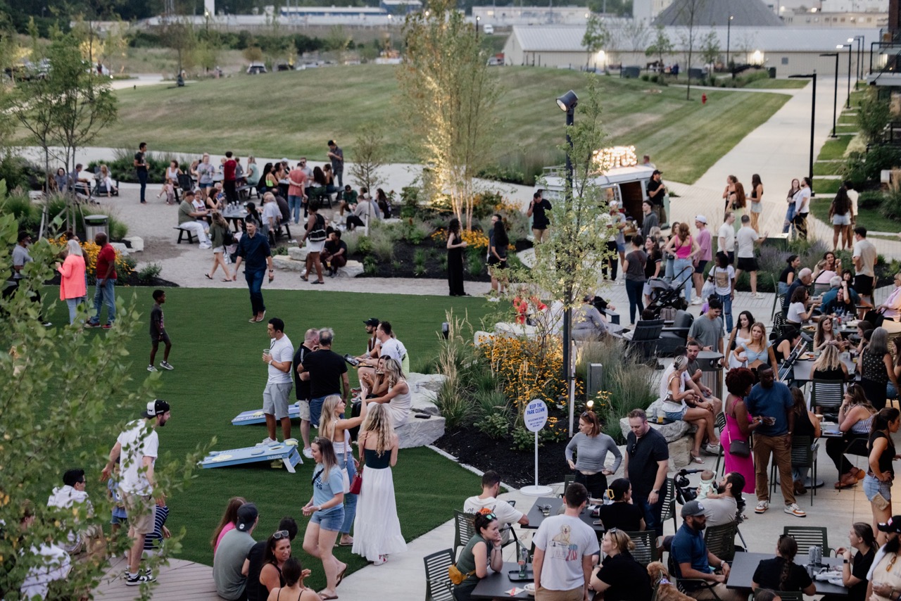 Outdoor gathering in a park with people socializing, sitting on grass and benches, and playing games. Trees and pathways are visible in the background.