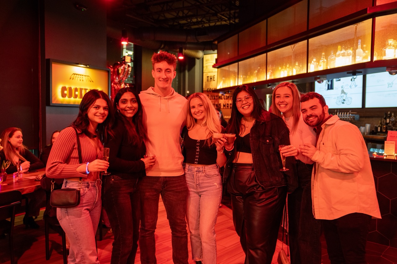 A group of seven people standing close together in a dimly lit restaurant, holding drinks and smiling at the camera.