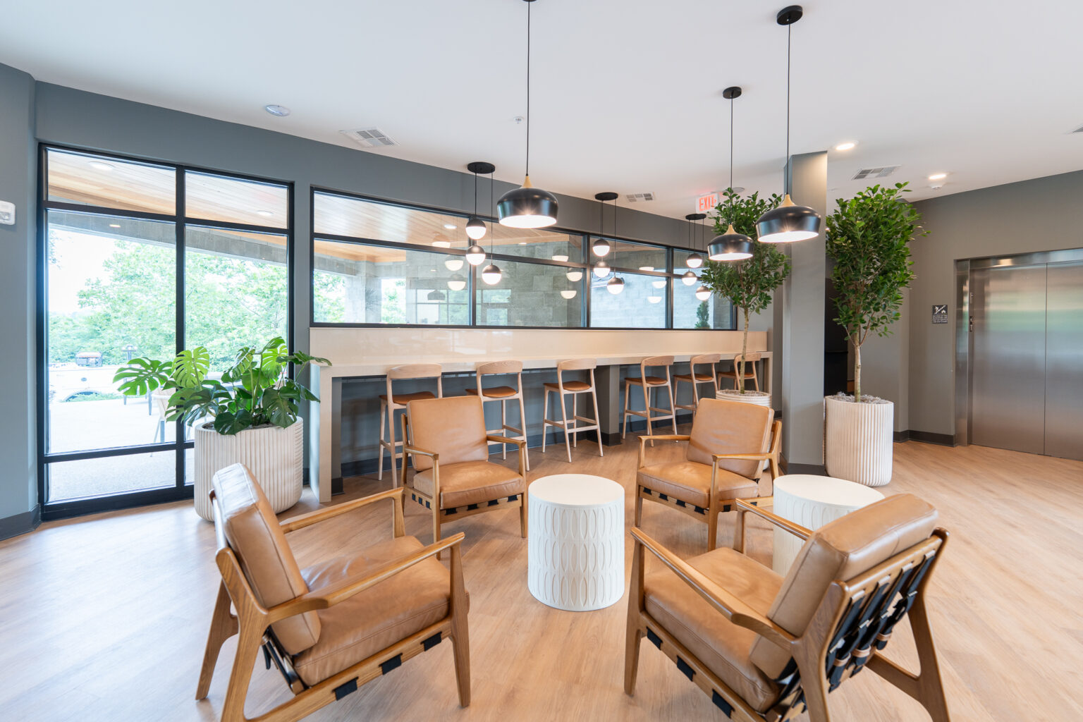 Modern lounge area with four tan leather chairs, two small white tables, a row of wooden stools at a counter, large windows, potted plants, and hanging lights.