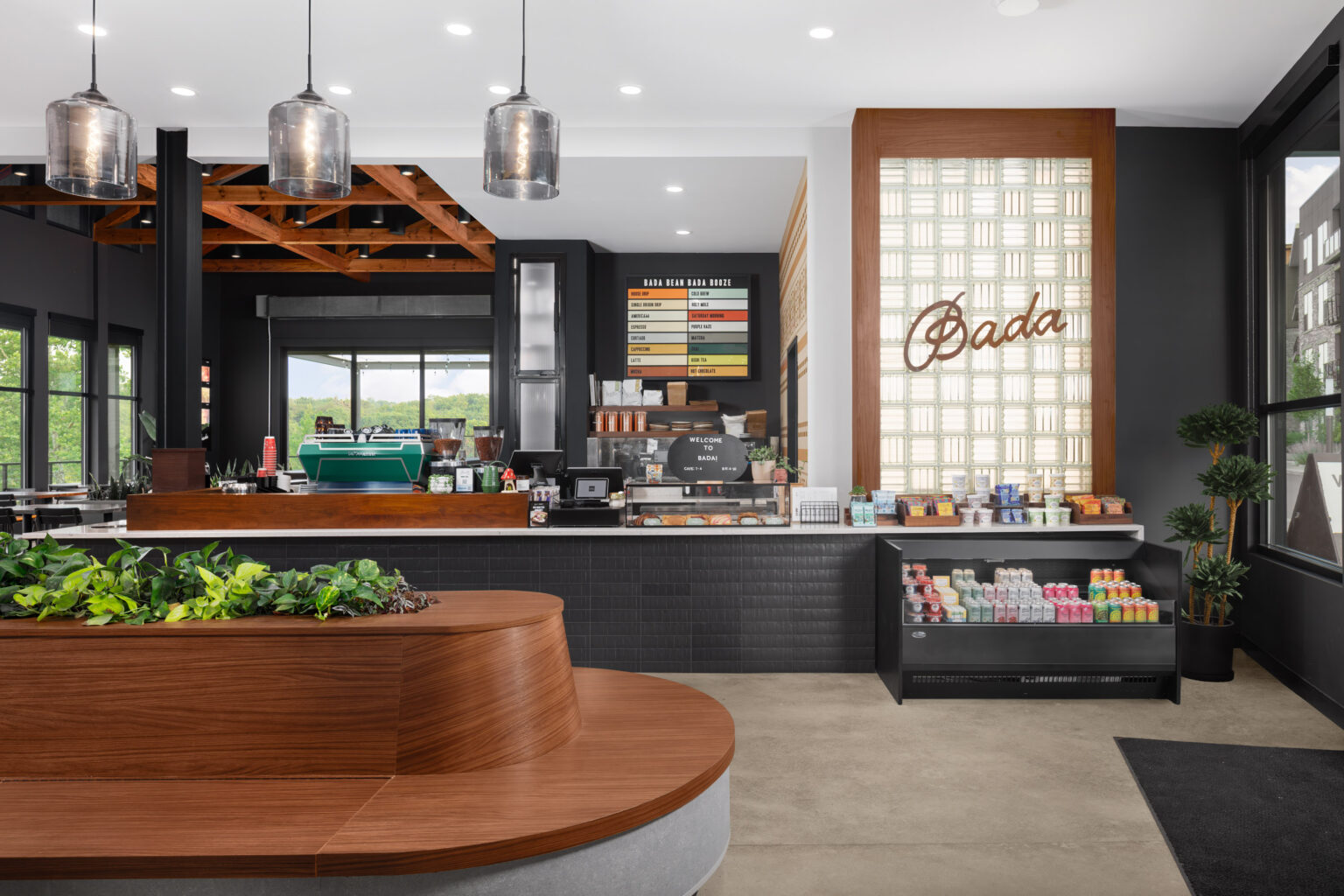 Modern coffee shop interior with a wooden counter, espresso machines, a pastry display, drink cooler, and a menu board on the wall.