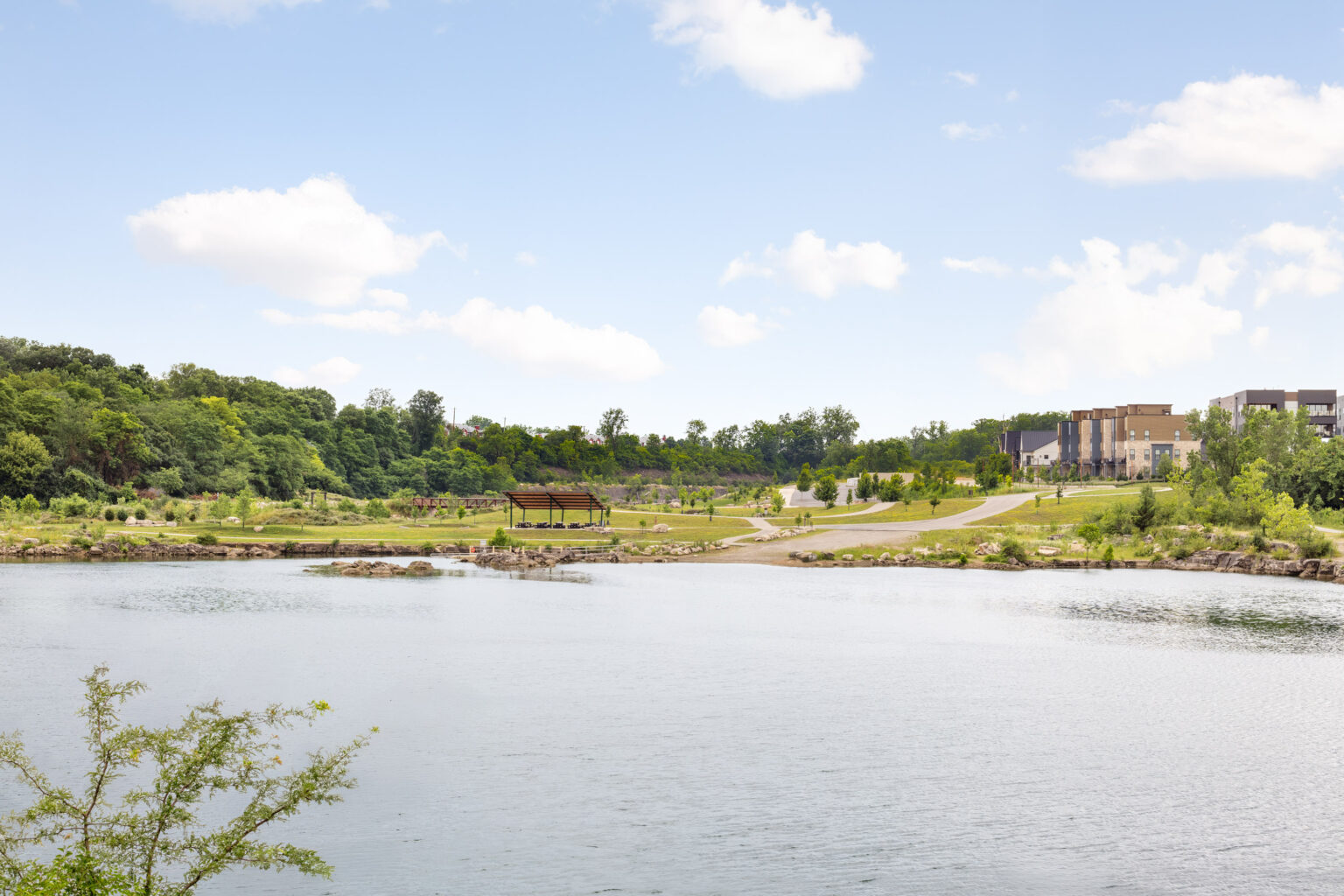 A calm lake with a small pavilion on the shore, grassy park areas, trees, and modern buildings in the background under a partly cloudy sky.