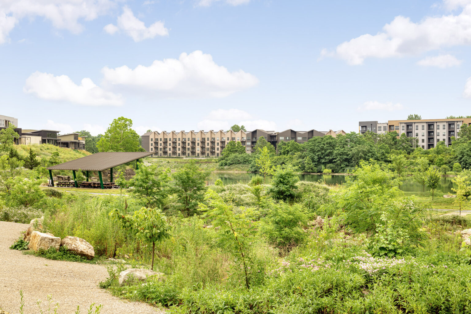 A landscaped park with a covered pavilion, gravel path, and greenery in the foreground, with modern apartment buildings in the background under a partly cloudy sky.