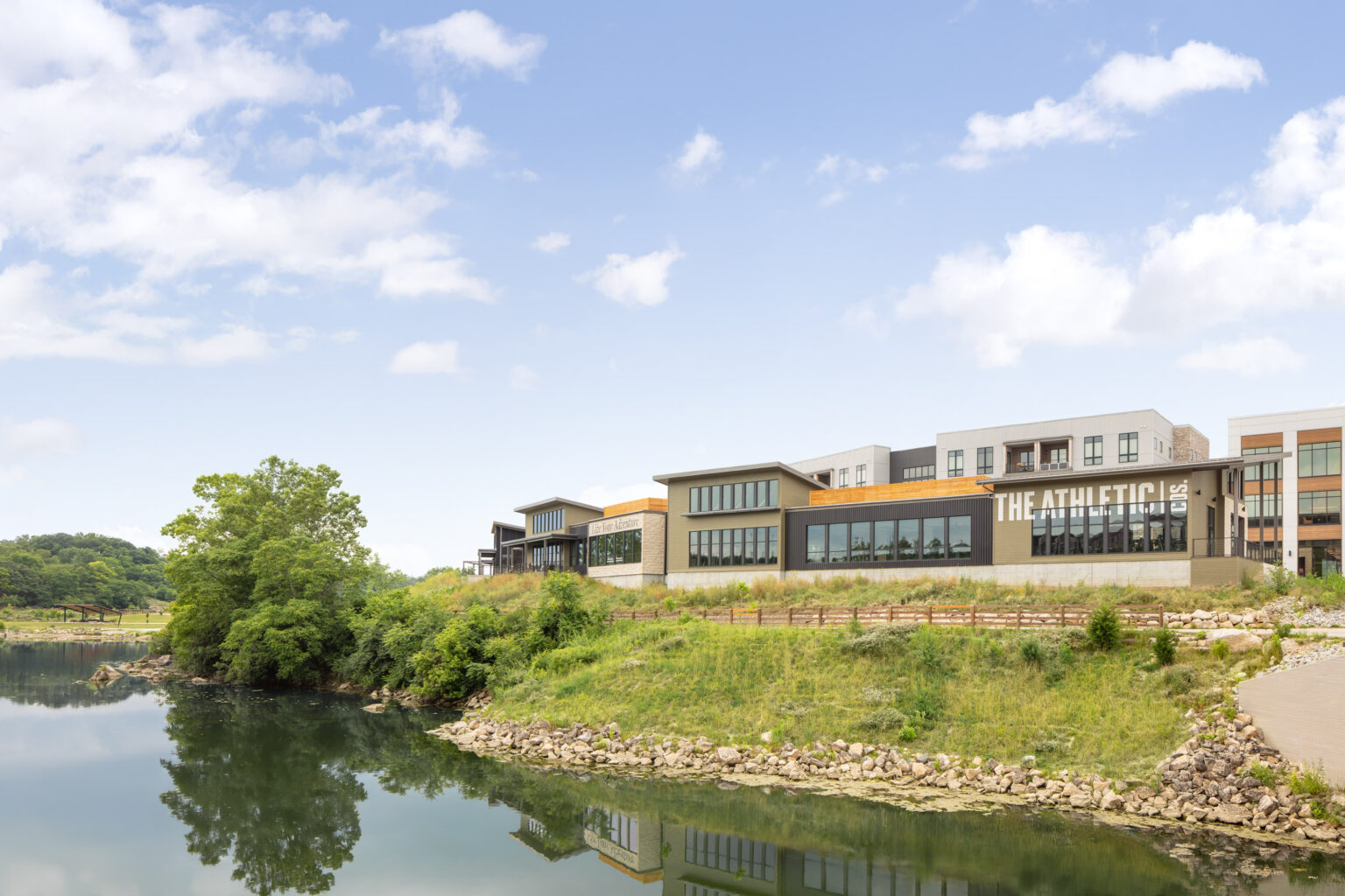 A modern building labeled “The Athletic” sits on a grassy riverbank with clear water, trees, and a partly cloudy sky in the background.