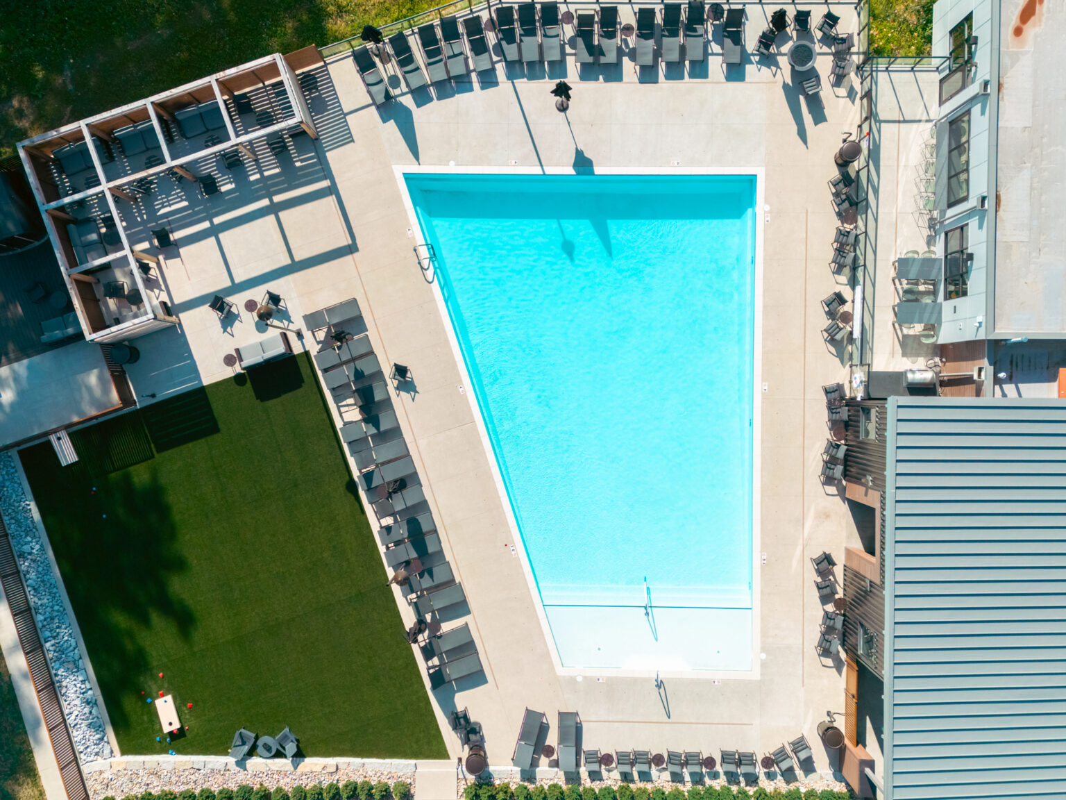Aerial view of a rectangular outdoor swimming pool surrounded by lounge chairs, artificial grass, and shaded seating areas.