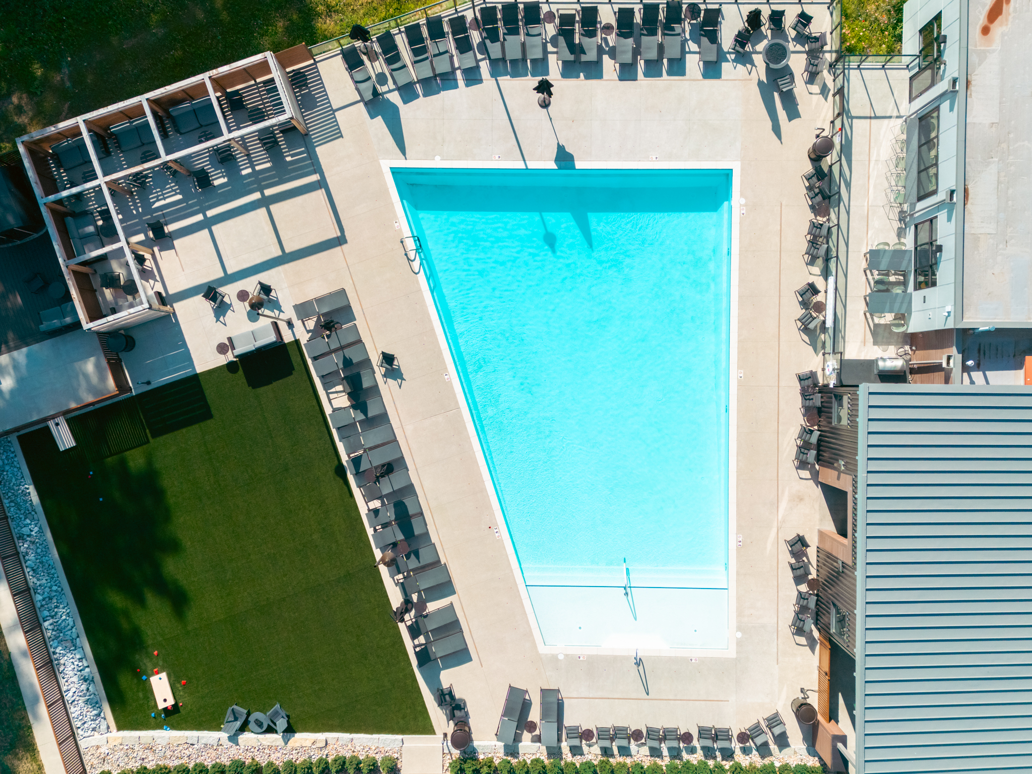 Aerial view of a rectangular outdoor swimming pool surrounded by lounge chairs, artificial grass, and shaded seating areas.