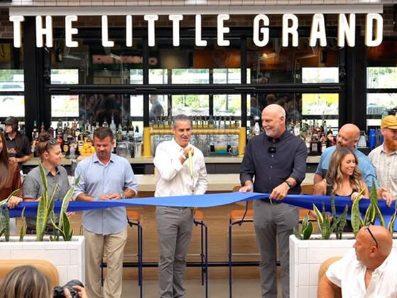 A group of people gathers for a ribbon-cutting ceremony at The Little Grand. A man in the center is about to cut the blue ribbon. The background features a bar with visible signage.
