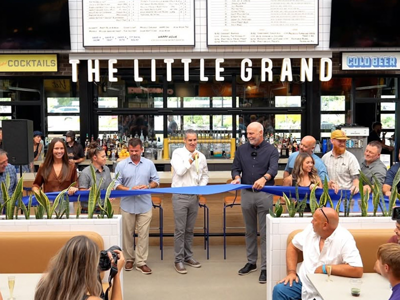 A group of people gathers for a ribbon-cutting ceremony at The Little Grand. A man in the center is about to cut the blue ribbon. The background features a bar with visible signage.