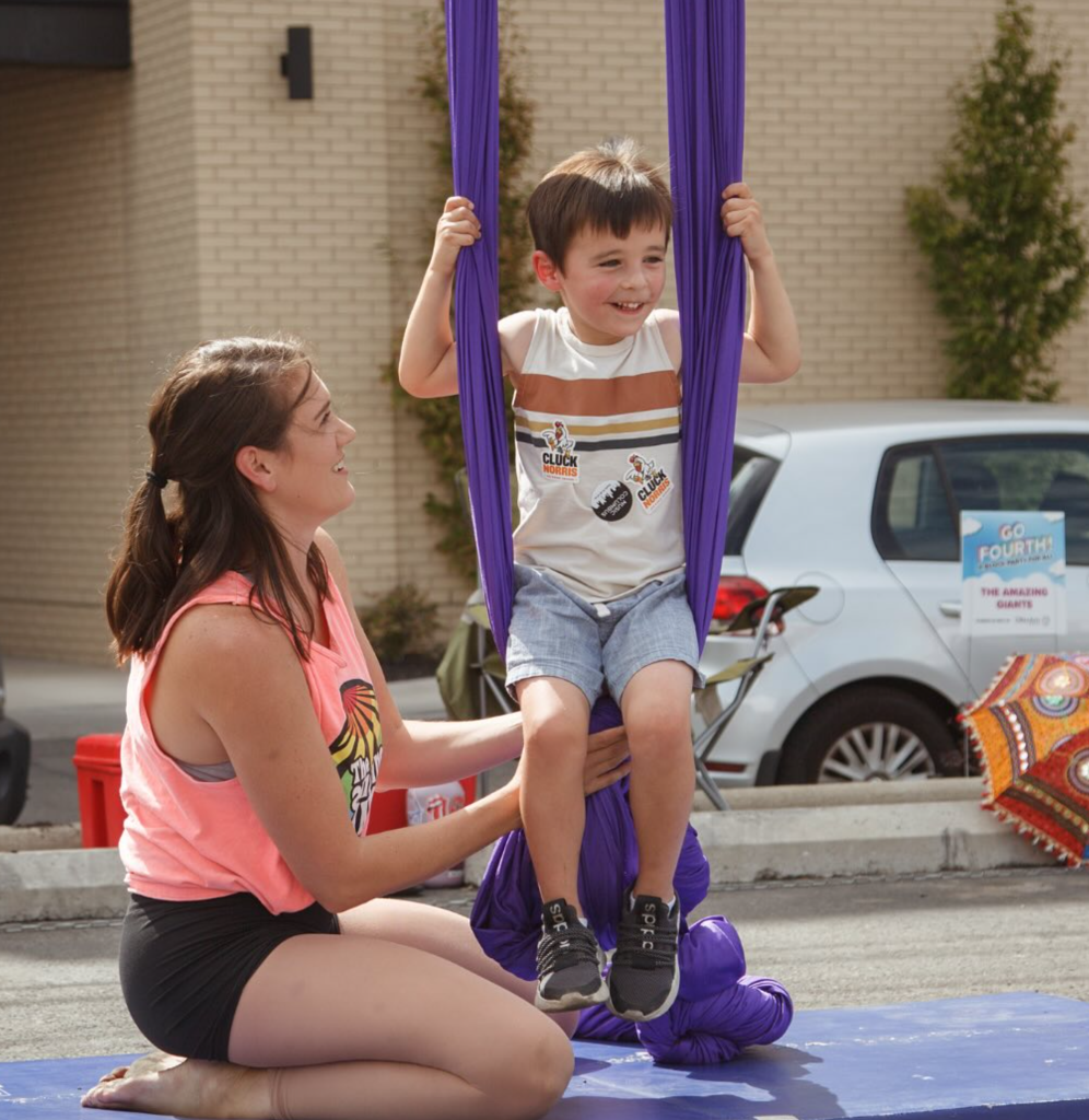 A woman helps a smiling young boy sitting in purple aerial silk during an outdoor event. A car and an umbrella are in the background.