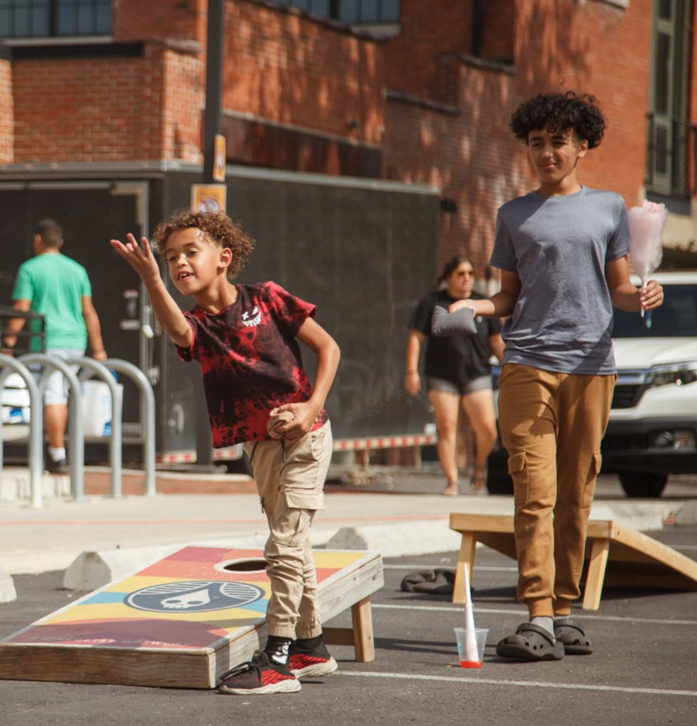 Two kids playing cornhole outdoors. One throws a beanbag, while the other holds cotton candy. Adults and a car are in the background.