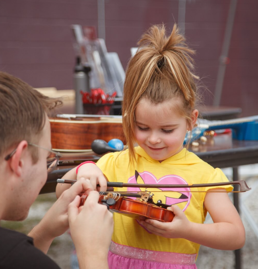 A young girl in a yellow shirt learns to play a small violin with guidance from a man, outdoors.