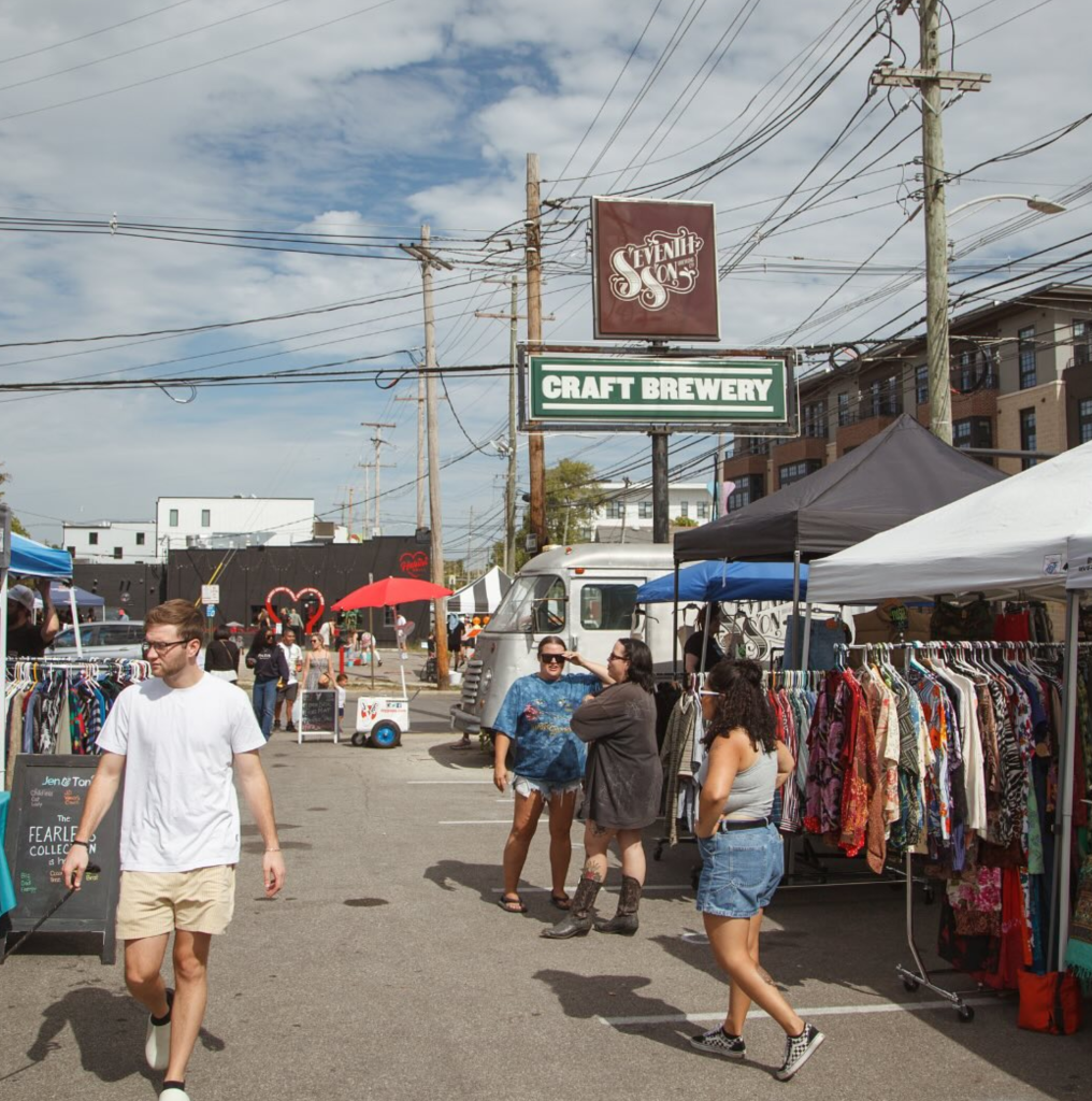 Outdoor market scene with people browsing clothing racks under tents. A sign reads Craft Brewery above a silver truck. Power lines and buildings are in the background.