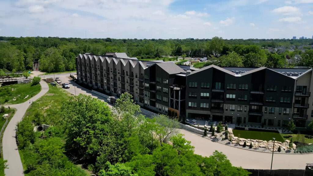 A modern four-story apartment building with a zigzag roofline is surrounded by trees, with a walking path and parking lot visible nearby under a partly cloudy sky.
