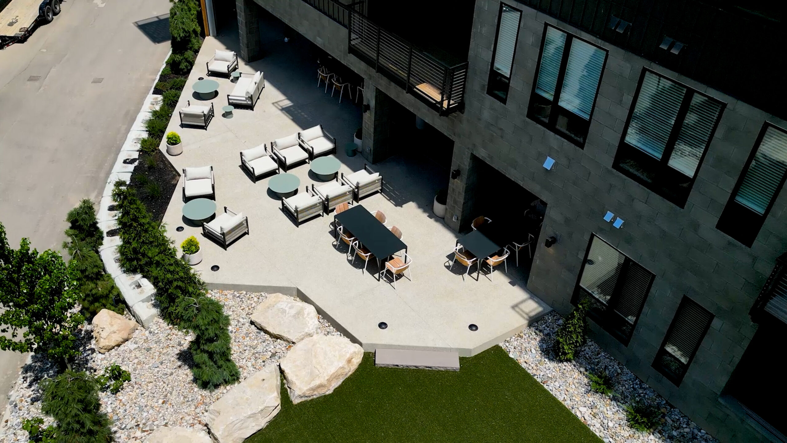 A modern outdoor patio with lounge chairs, round tables, and a long dining table is adjacent to a building, bordered by landscaping with rocks and greenery.