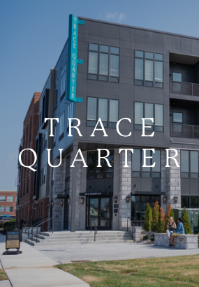 A modern, multi-story apartment building named Trace Quarter stands tall, with its sign on the facade and two people relaxing on benches near the entrance.